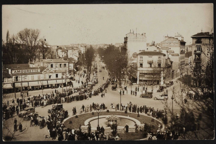 s.t, Boulogne-Billancourt . avenue de la Reine, rond-point Rhin-et-Danube . - PerspectiveVue plongeante sur le rond-point, au premier plan et l'avenue de la Reine, au second plan. Cortège d'automobiles remontant l'avenu vers le rond-point. Une foule les regarde passer. Au second plan, à gauche, le café Damourette et une pâtisserie.