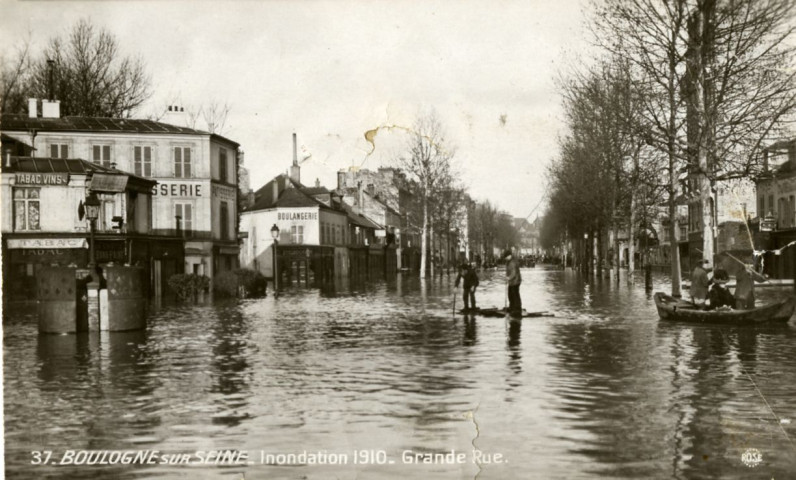 Boulogne-sur-Seine - Inondation 1910 - Grande-Rue, Boulogne-Billancourt . avenue Jean-Baptiste Clément . - Crue de la Seine, janvier 1910 - l'avenue Jean-Baptiste Clément inondée.- A gauche, un café "La civette du rond point", une pâtisserie et une boulangerie. Au premier plan, au centre et à droite des personnes sur des embarcations. Au fond, un groupe regarde la scène.